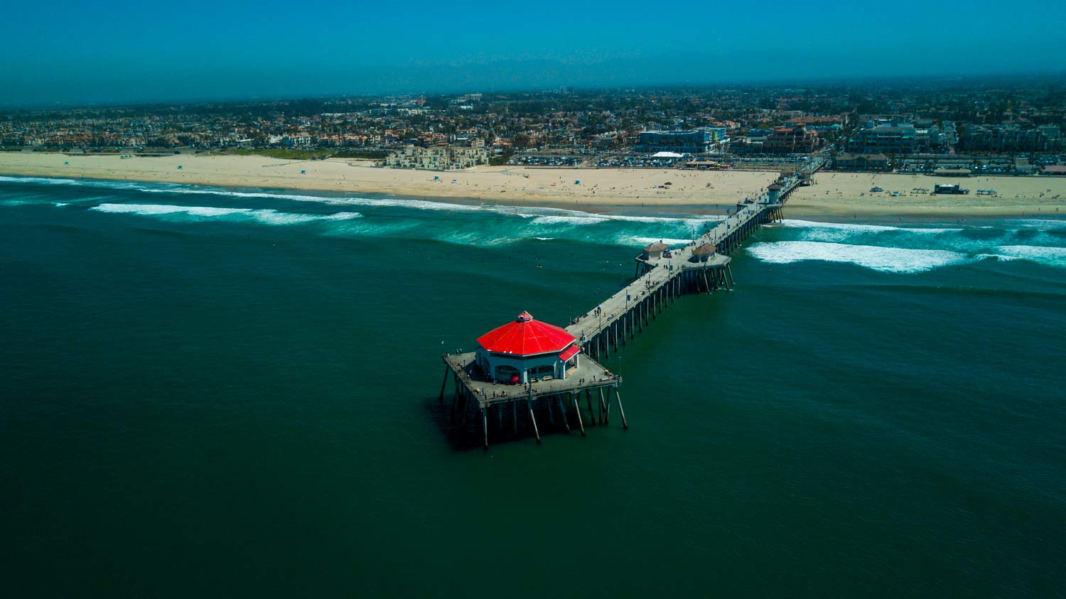 Aerial view of a pier with a red-roofed structure extending into turquoise ocean beside a sandy beach.