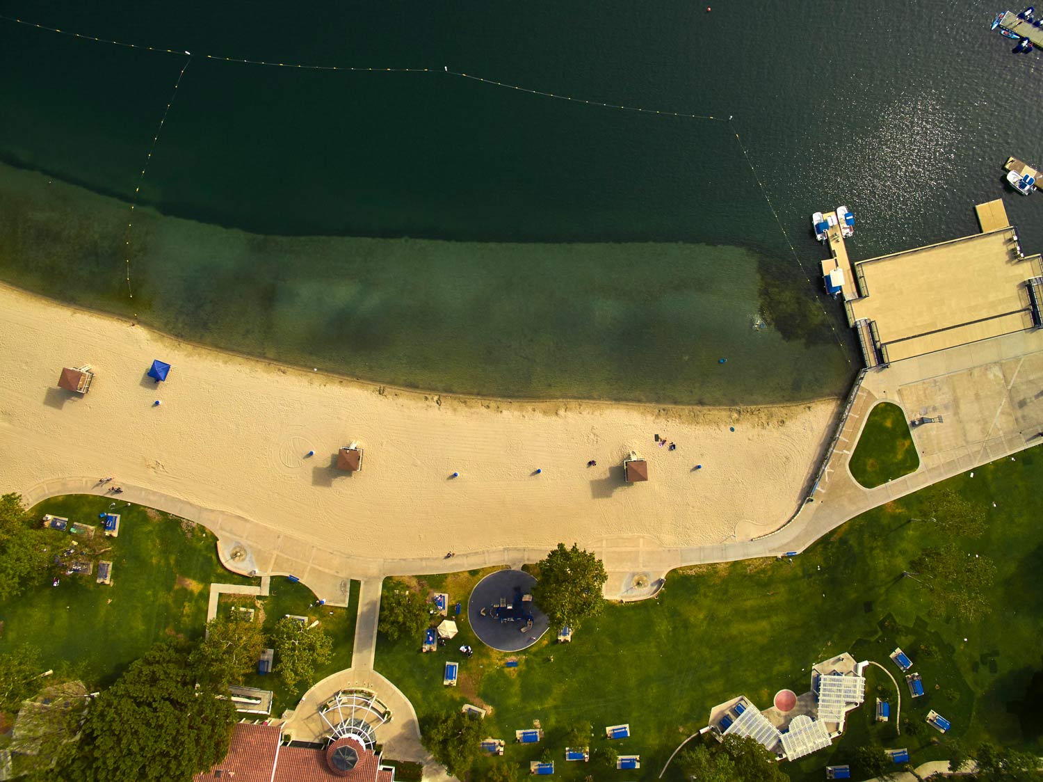 Aerial view of a sandy beach beside dark water, with a wooden dock and a small grassy area nearby.
