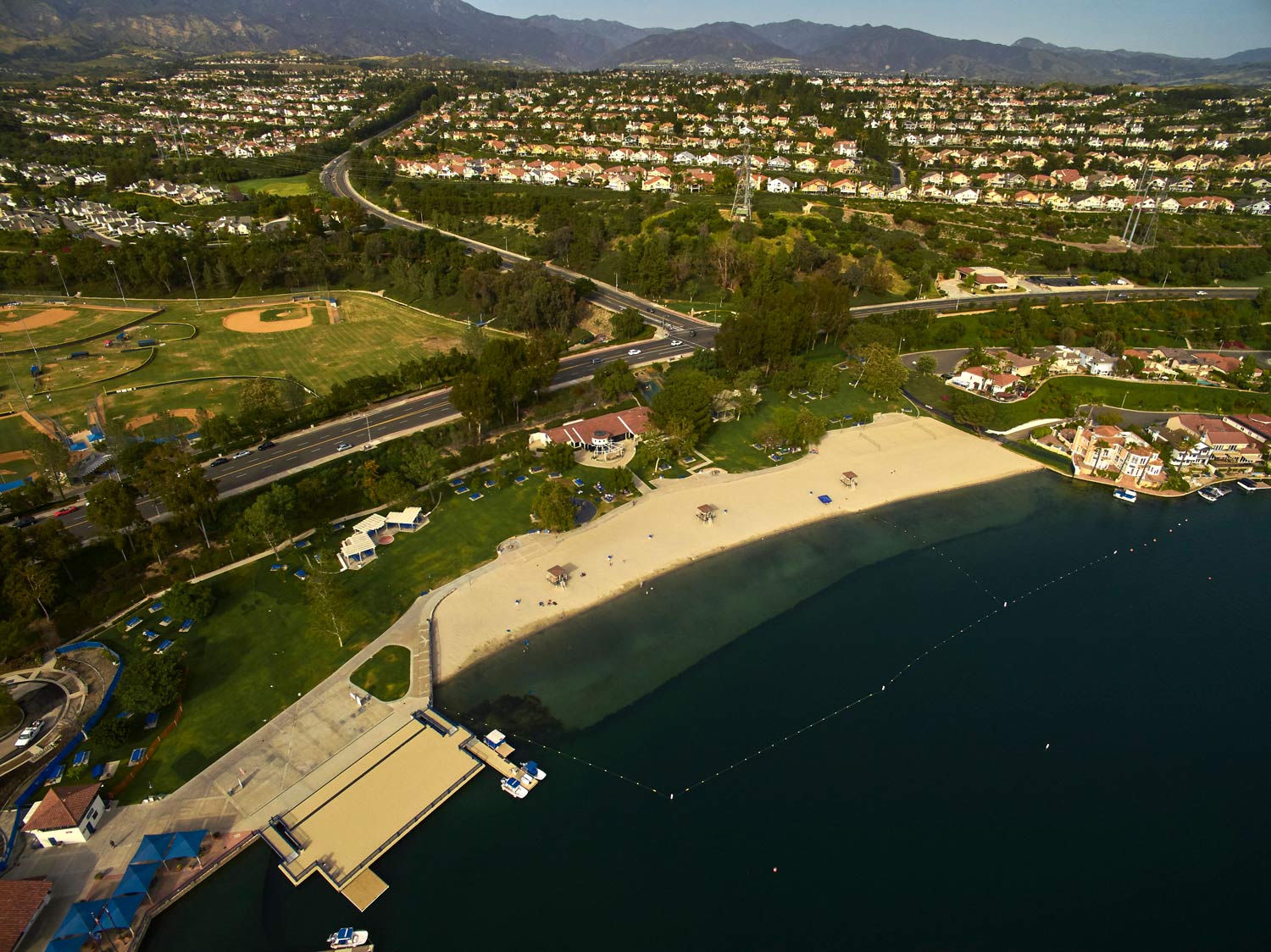 Aerial view of a sandy beach, pier, and coastal town beside dark blue water and hills.
