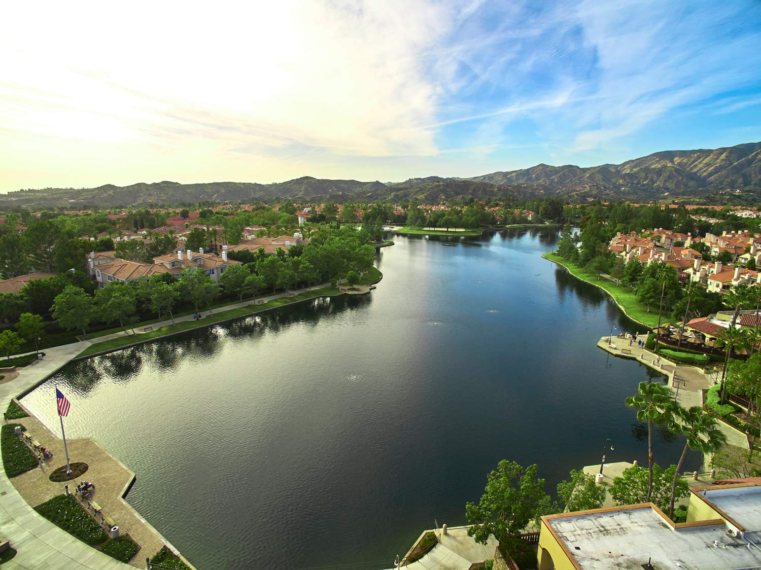 Aerial view of a calm lake in a green residential area, with hills and a partly cloudy sky.