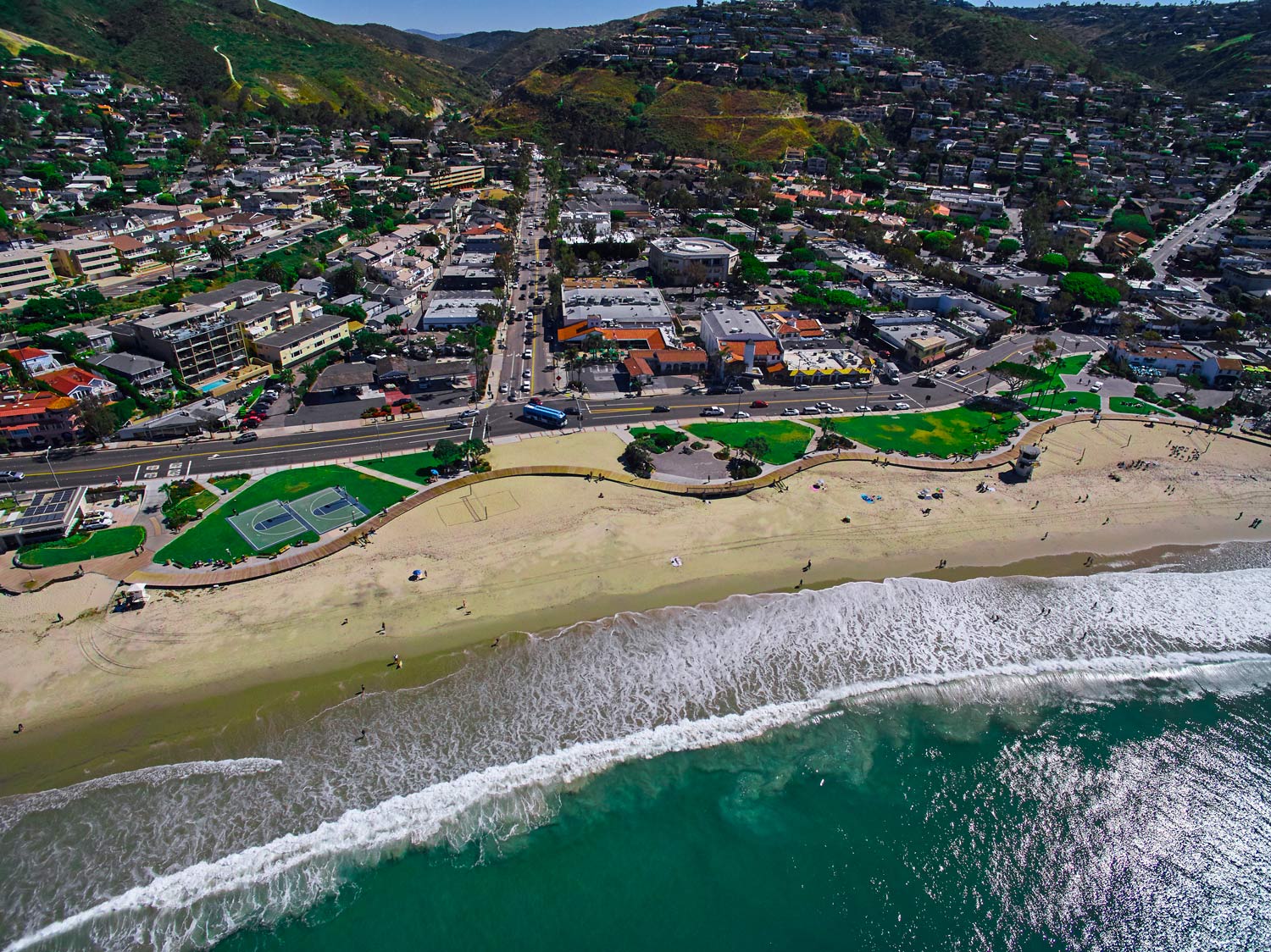 Aerial view of a coastal town with a beach, waves, and green hills behind it
