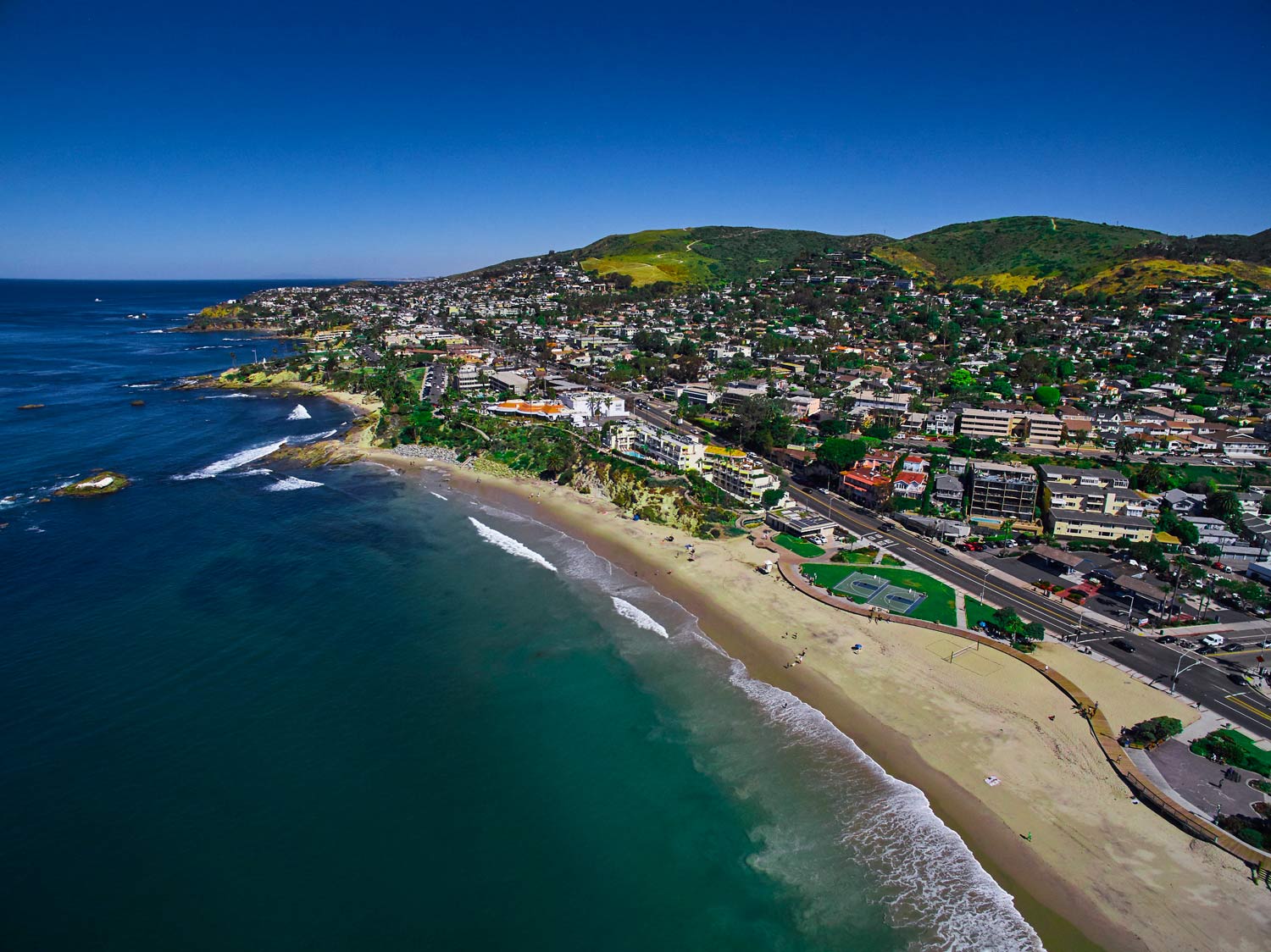 Aerial view of a coastal town beside a sandy beach and blue ocean under green hills