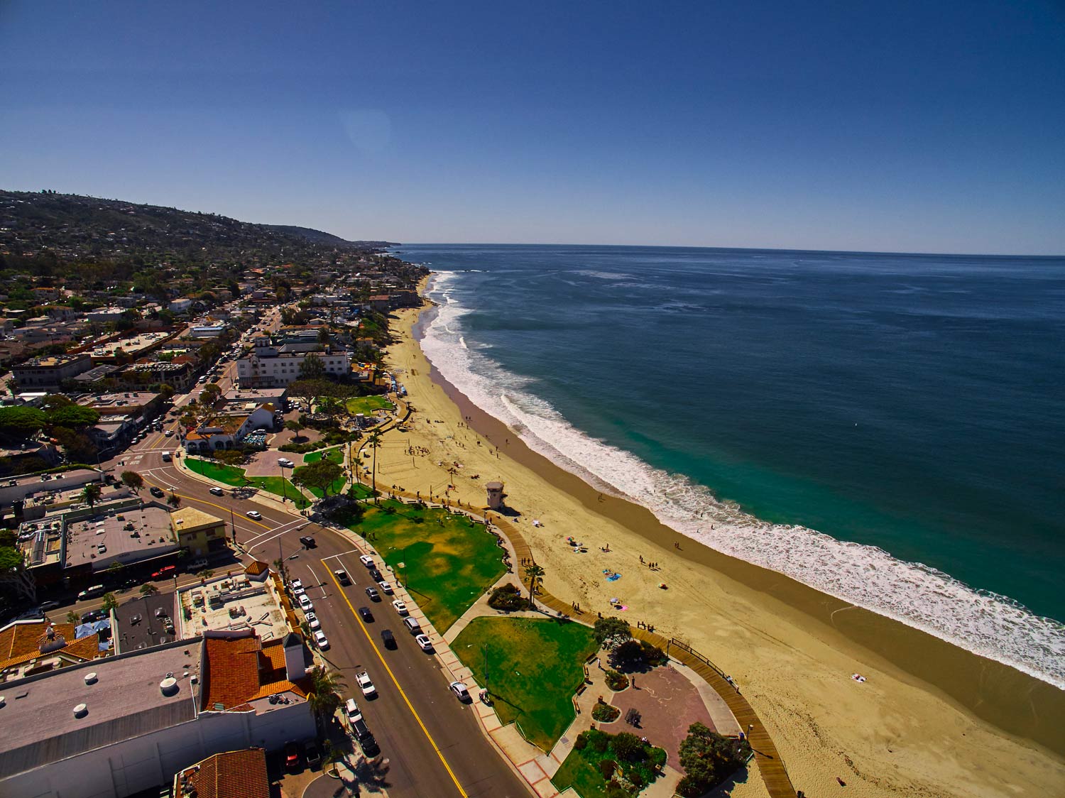 Aerial view of a sandy beach beside a coastal road, ocean waves, and hillside buildings under a clear blue sky