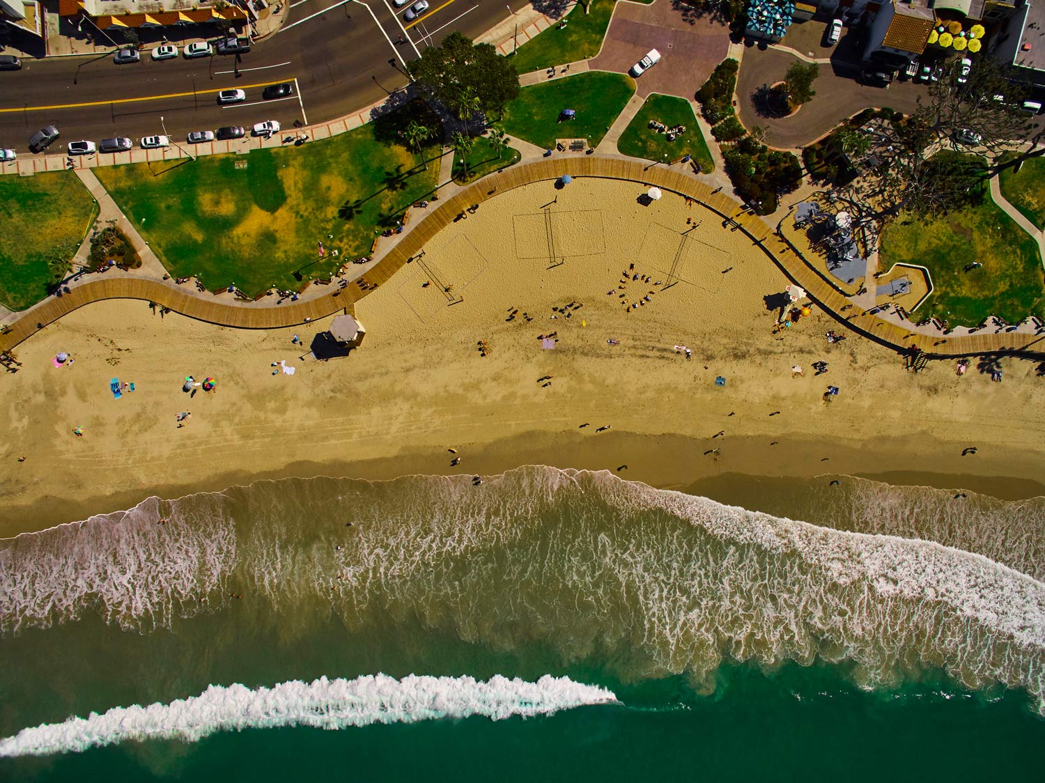 Aerial view of a sandy beach with waves, people, and a road lined with trees above.