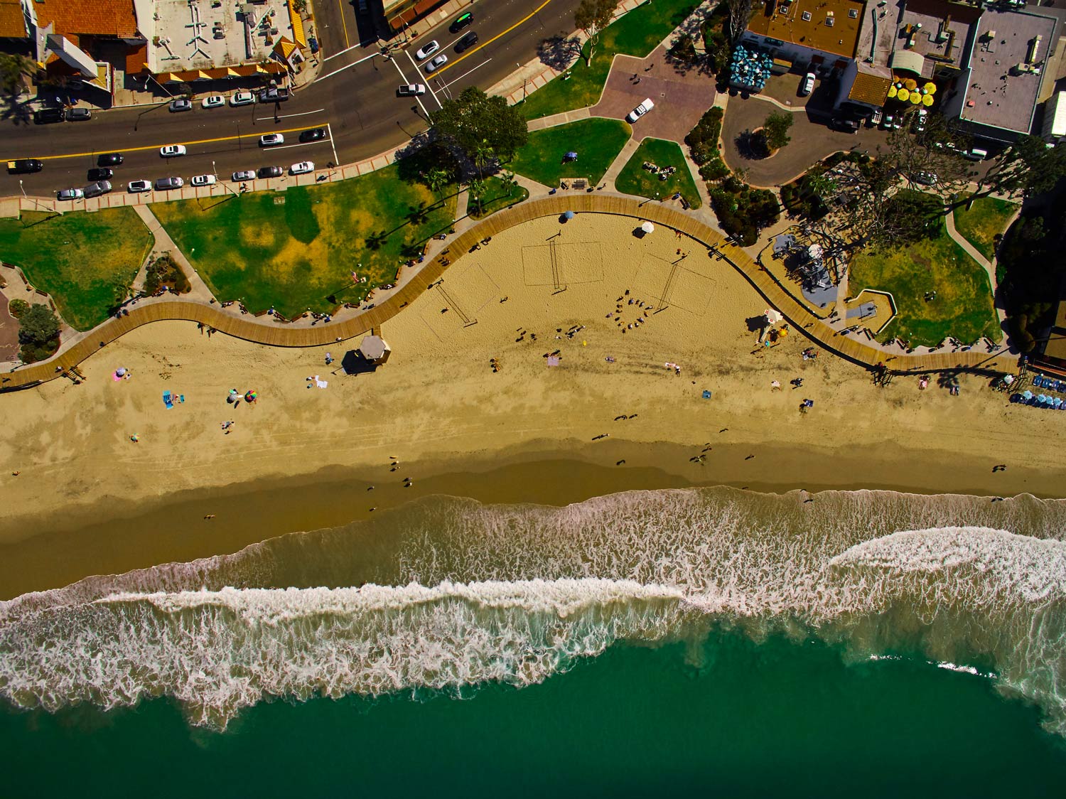 Aerial view of a sandy beach with waves, green water, and a walkway beside buildings and trees