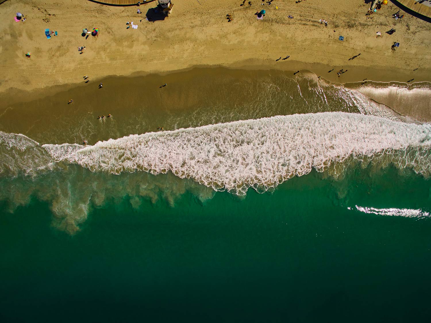Aerial view of a sandy beach with waves breaking into green water and tiny beachgoers scattered on shore
