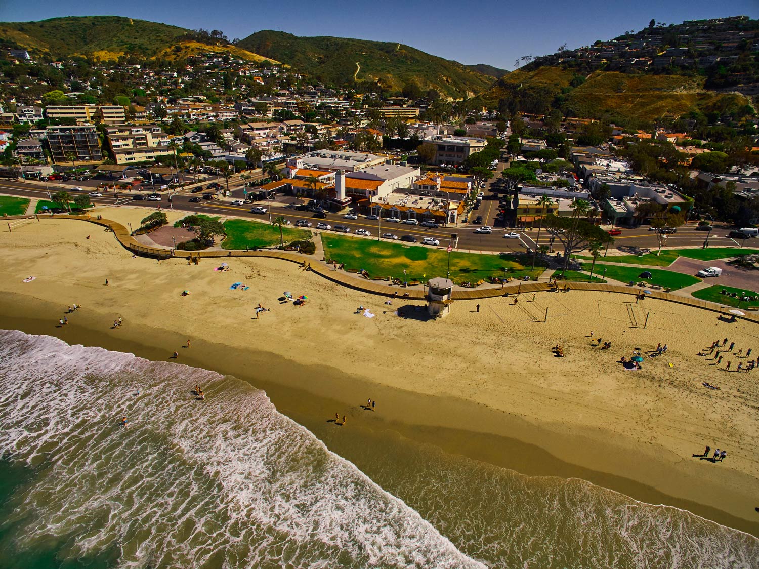 Aerial view of a sandy beach and surf beside a coastal town with green hills in the background