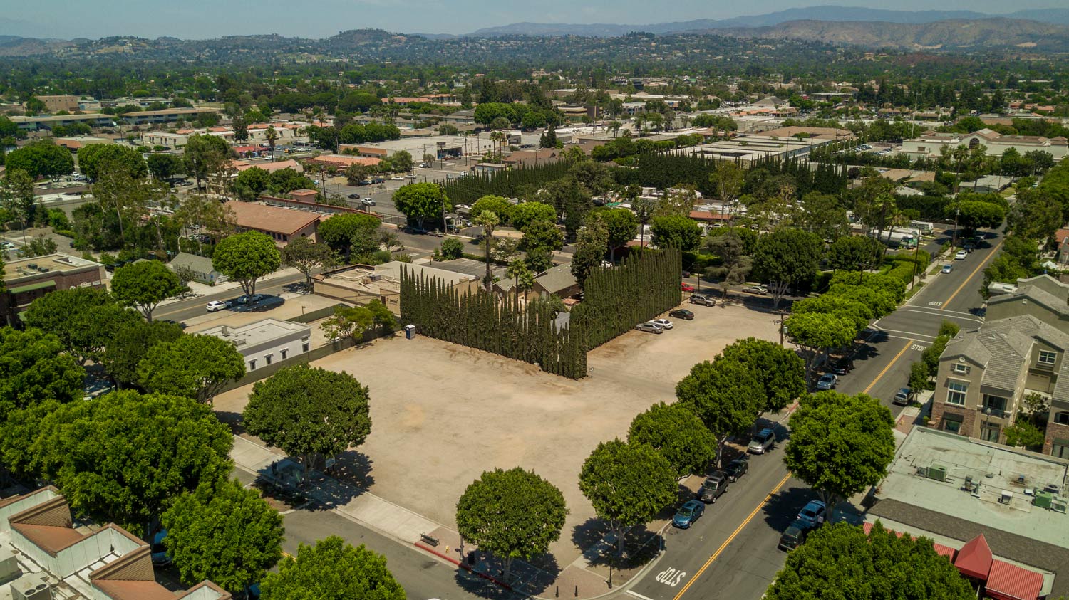 Aerial view of a leafy neighborhood with a large open dirt field and surrounding streets under clear skies