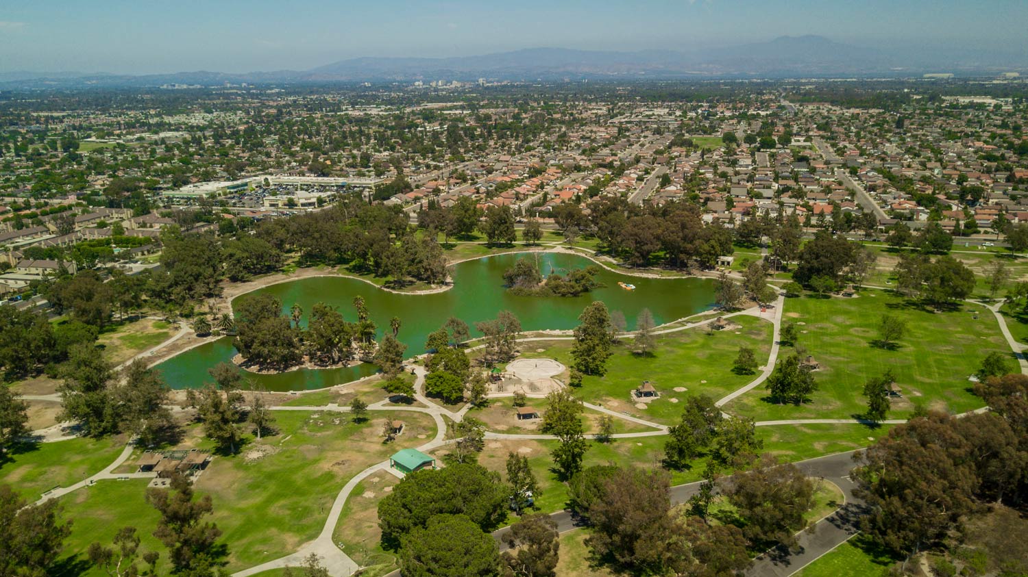 Aerial view of a green park with ponds and walking paths, surrounded by houses and city skyline.
