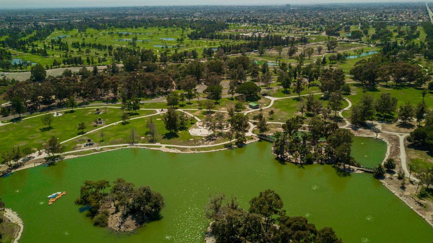 Aerial view of a park with a green lake, trees, walking paths, and surrounding cityscape