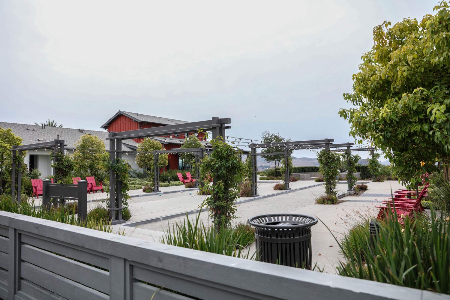Modern courtyard with gray paving, red-roofed building, pergola, and potted greenery under a cloudy sky
