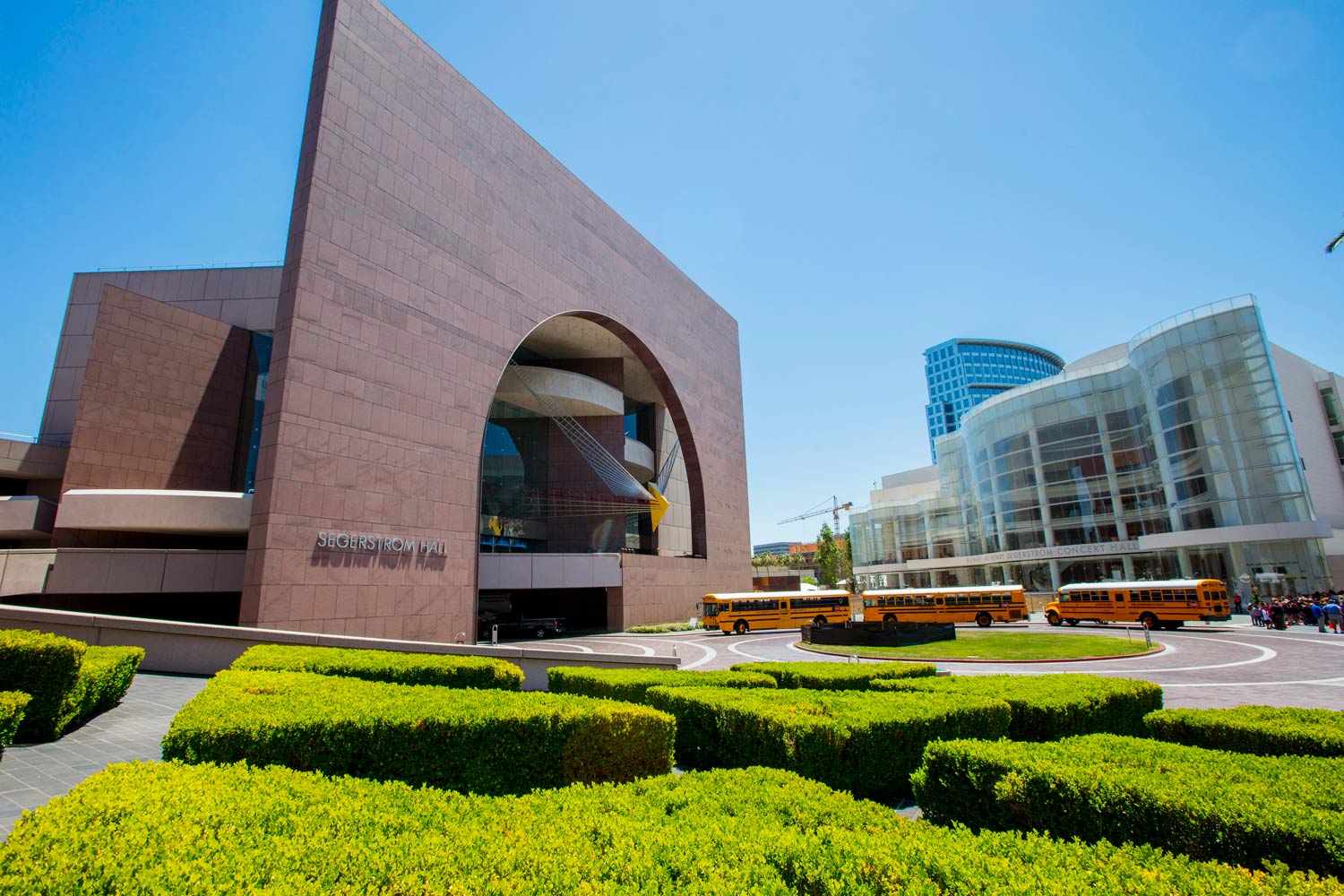Modern brick building with large arch and landscaped hedges beside a glass office tower under a blue sky