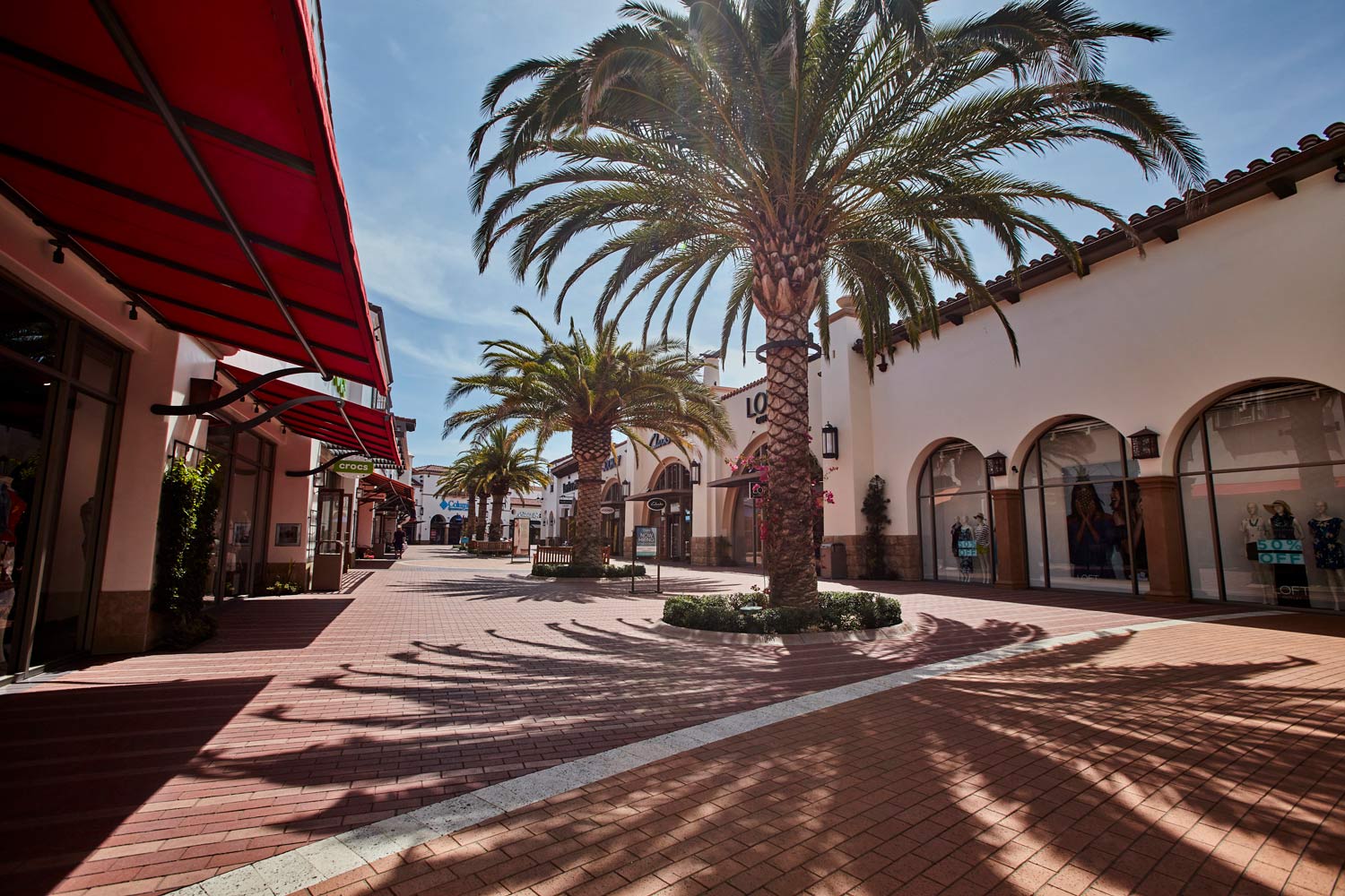 Sunny outdoor shopping plaza with palm trees, red awnings, and white stucco storefronts