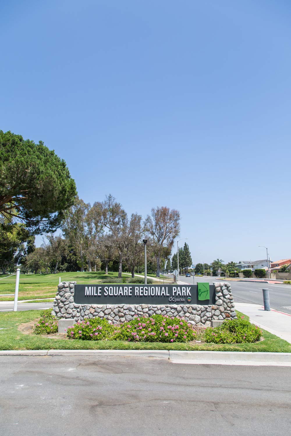 Mile Square Regional Park entrance sign beside a road, with trees, shrubs, and a clear blue sky.