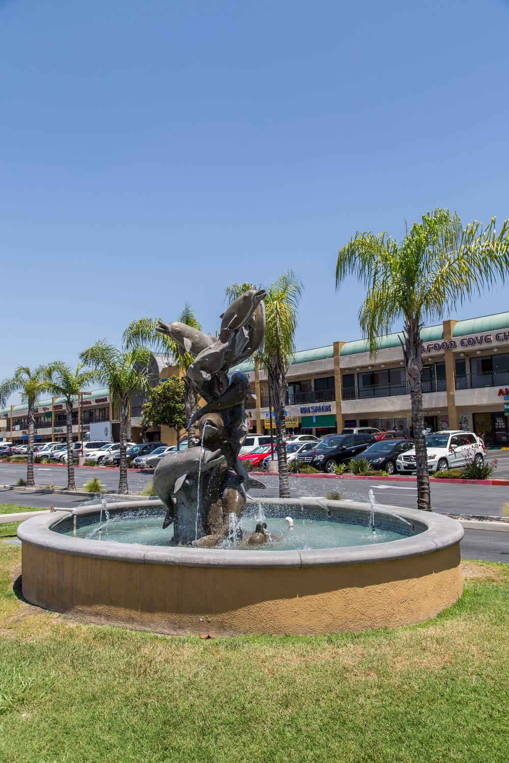 Decorative fountain with a twisting sculpture in a sunny parking lot beside palm trees and a shopping plaza