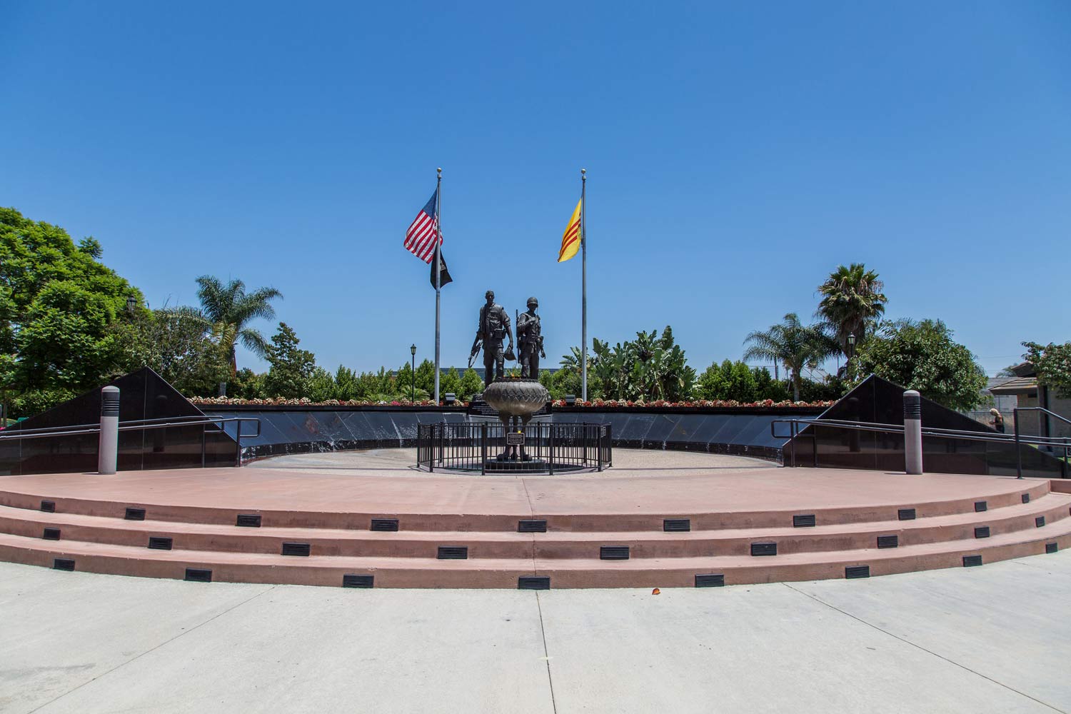 Memorial plaza with steps, two statues, and American and state flags under a blue sky