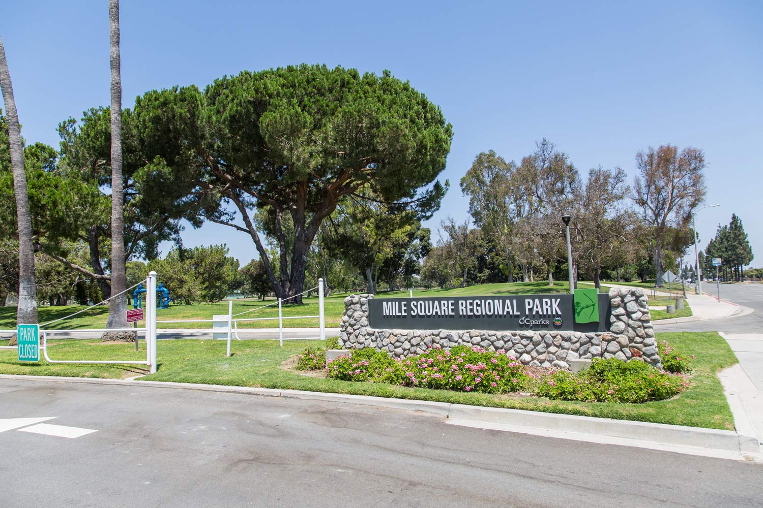 Entrance sign for a park with trimmed trees, grass, and a paved road under a clear blue sky
