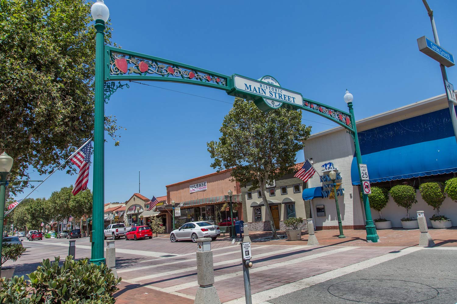 Decorative archway over a small-town street with storefronts, trees, and parked cars on a sunny day