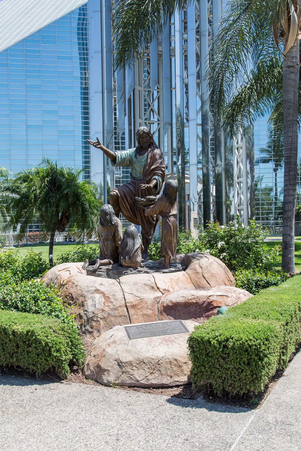 Bronze seated statue on a rock base in a landscaped plaza beside a glass office building.