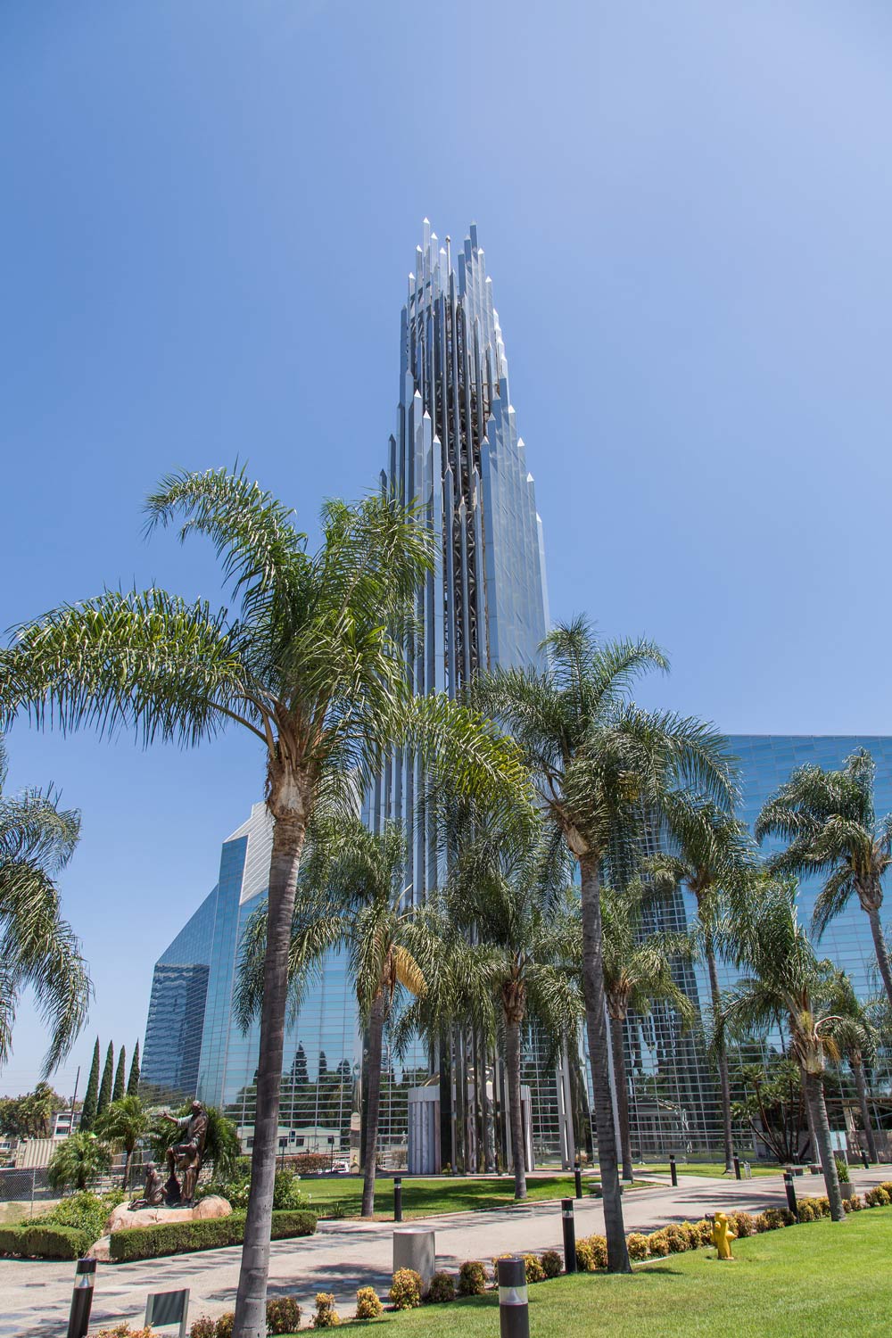 Tall glass skyscraper rising above palm trees in a sunny urban plaza.