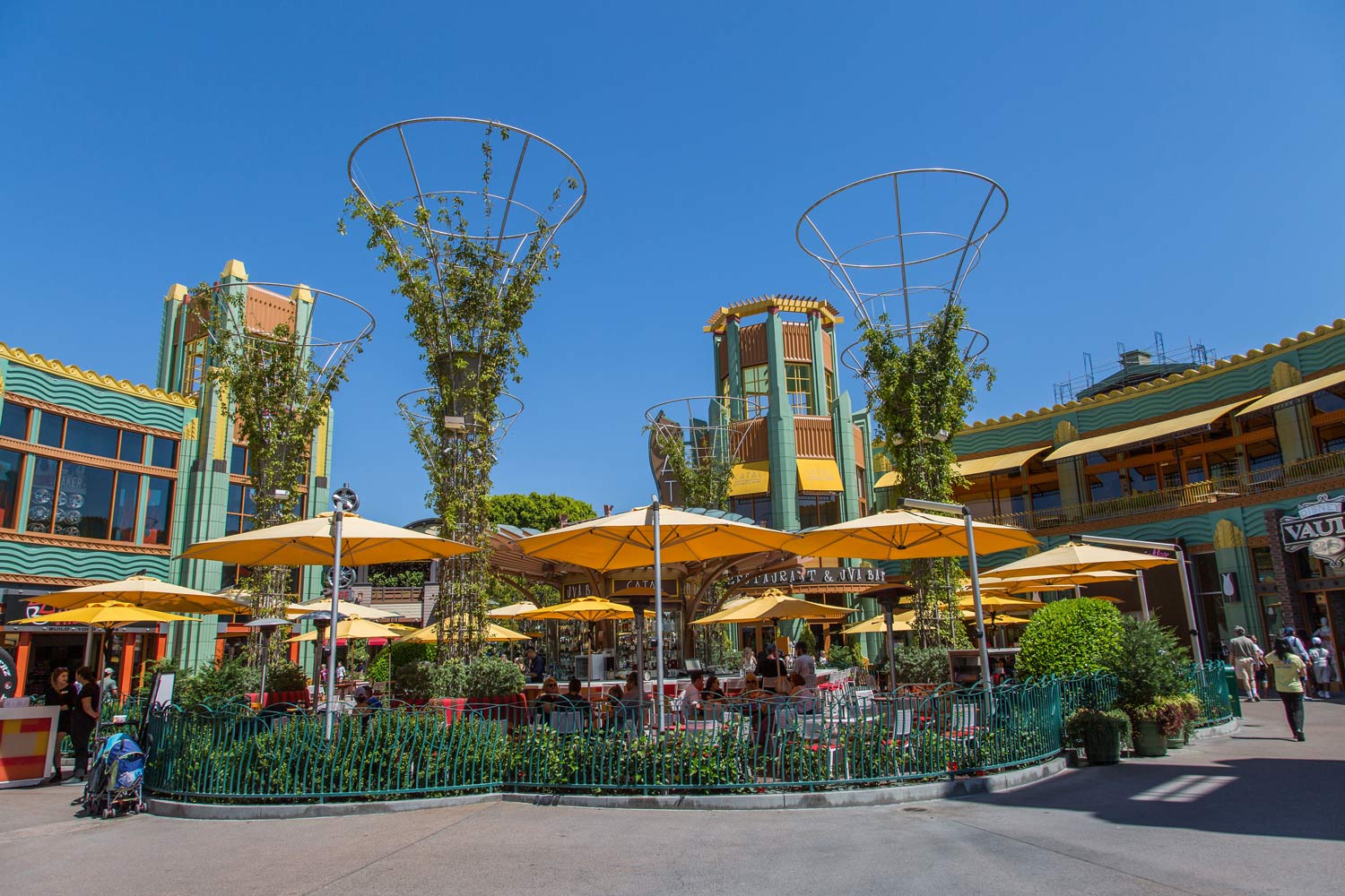 Outdoor plaza with colorful umbrellas, greenery, and decorative metal tree sculptures under a blue sky