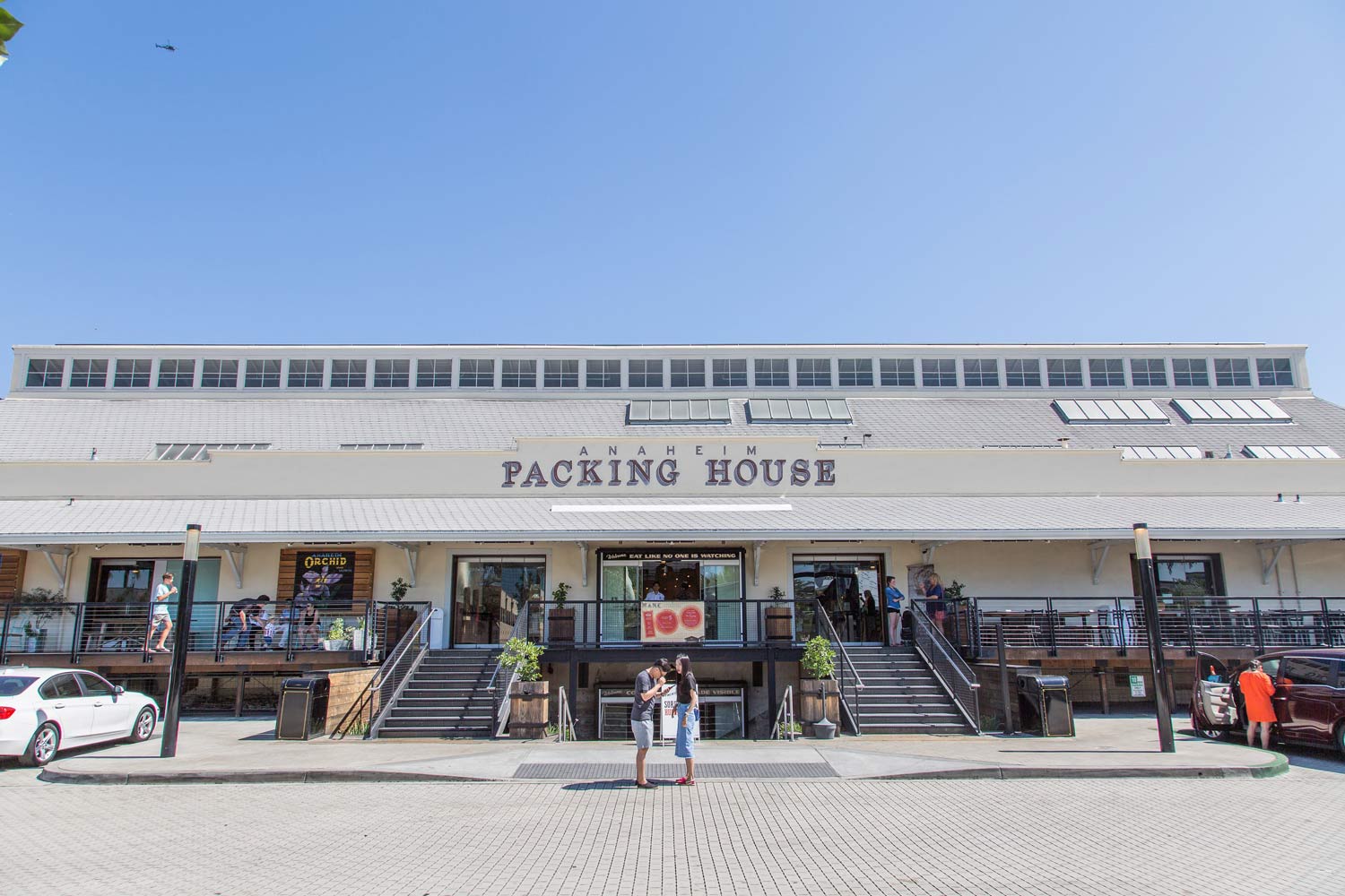 Front view of a “Packing House” building with people on the steps and parked cars in front