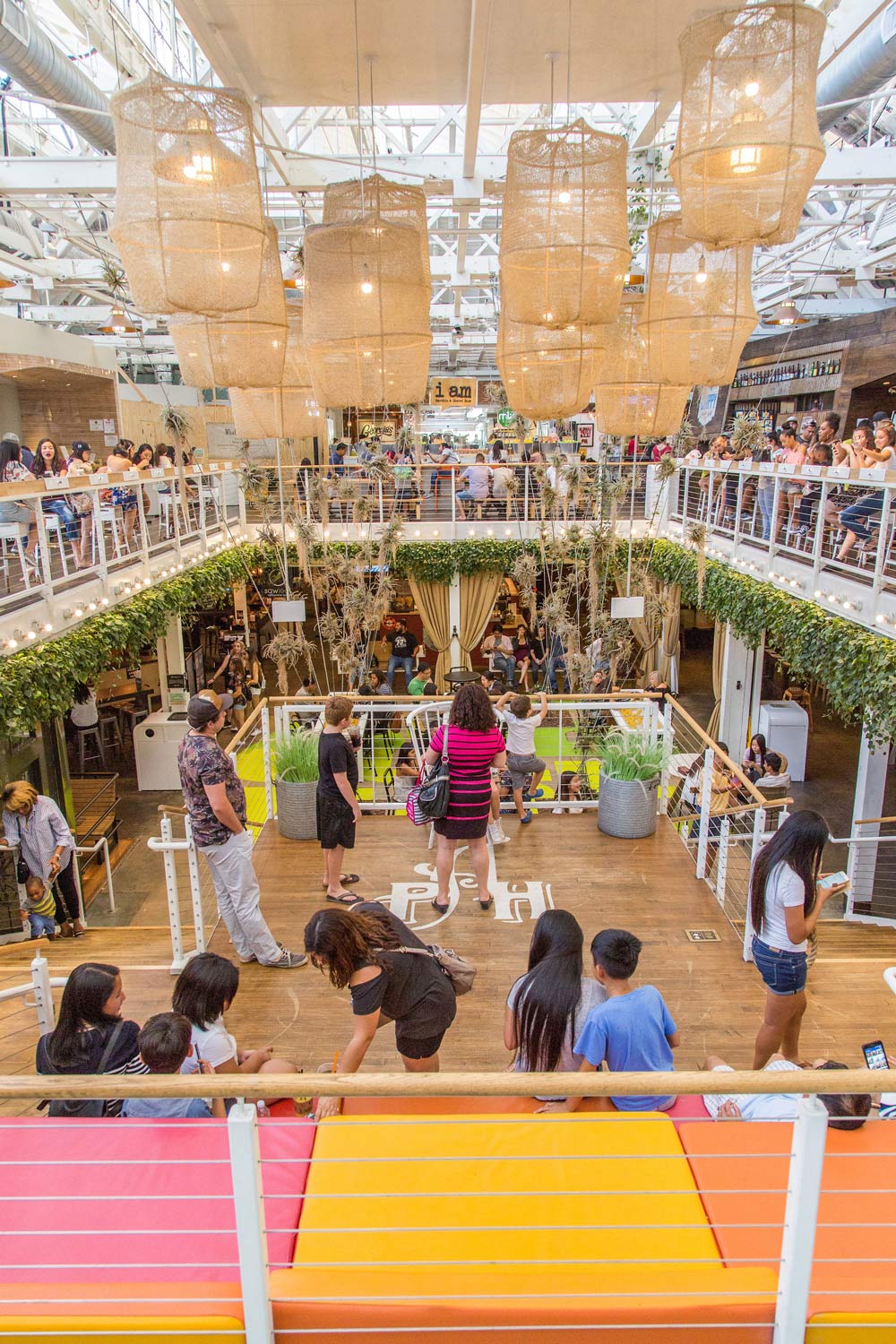 Bustling indoor atrium with hanging wicker lanterns, lush plants, and people gathered around a central walkway.