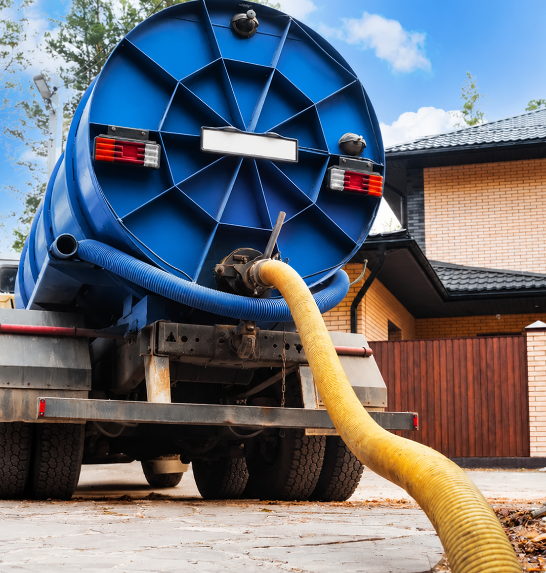 Blue vacuum truck with a yellow hose on a driveway outside a house.