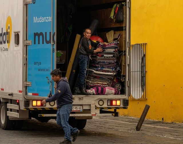 Una persona descarga mercancías apiladas de un camión junto a un edificio amarillo.