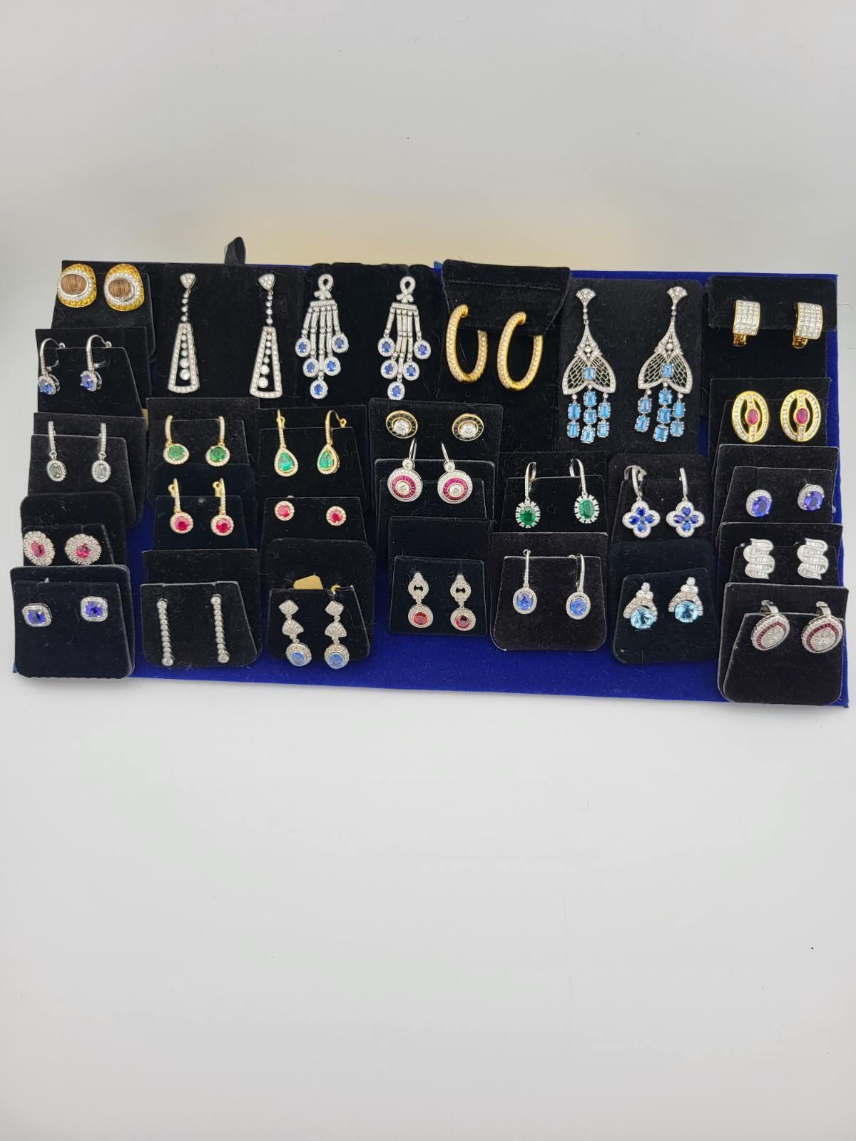 Display of various earrings on a blue velvet backdrop, including hoops, dangles, and studs, with gemstones.