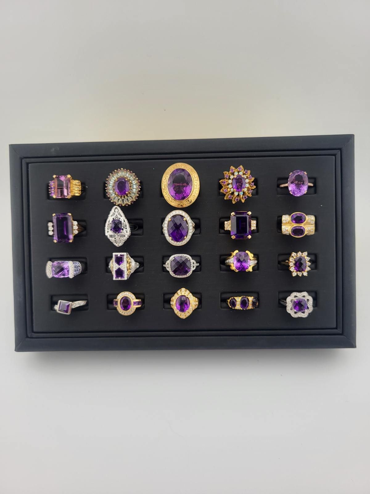 Tray of 20 amethyst rings with various settings on a black display surface.