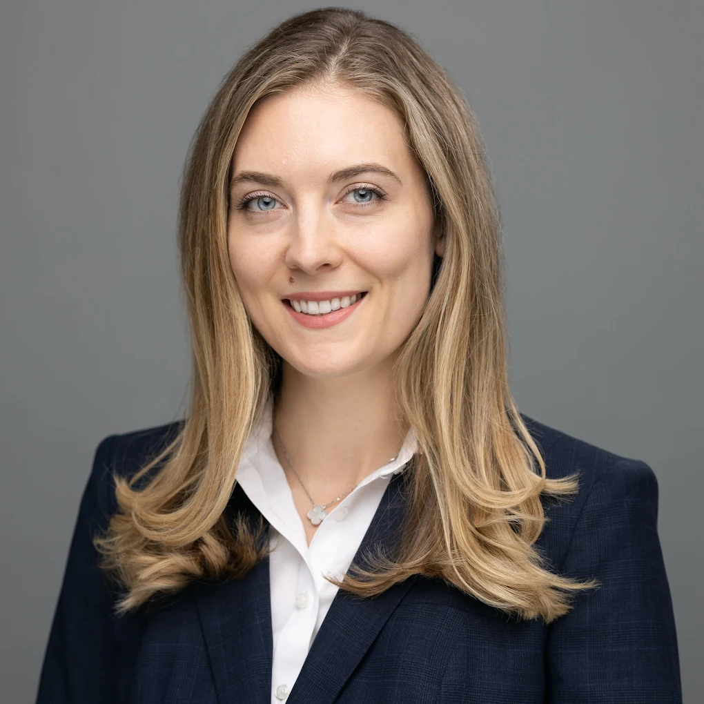 Woman with blonde hair wearing a white shirt and navy blazer smiles against a gray backdrop.