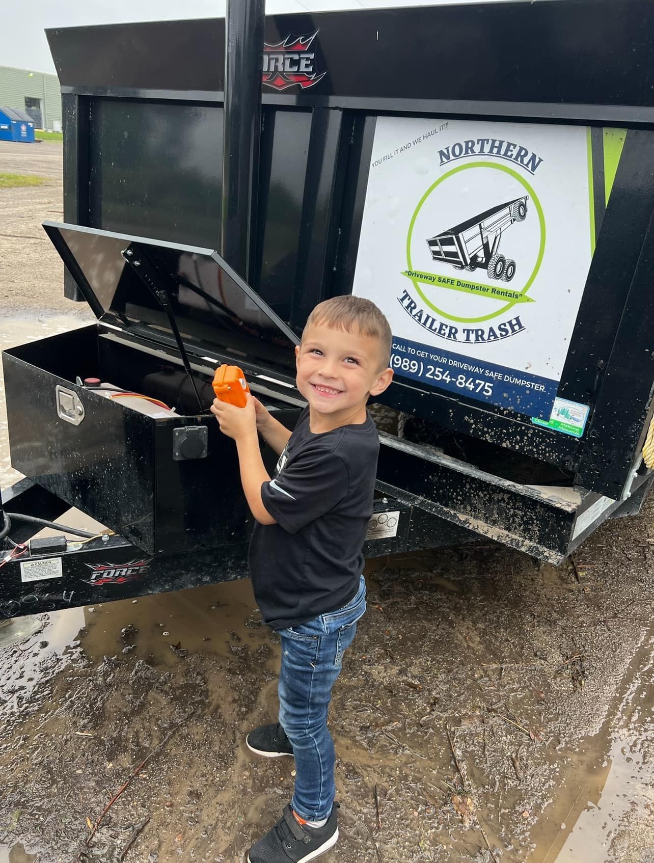 Young child smiling, holding orange object, beside a black trailer with 