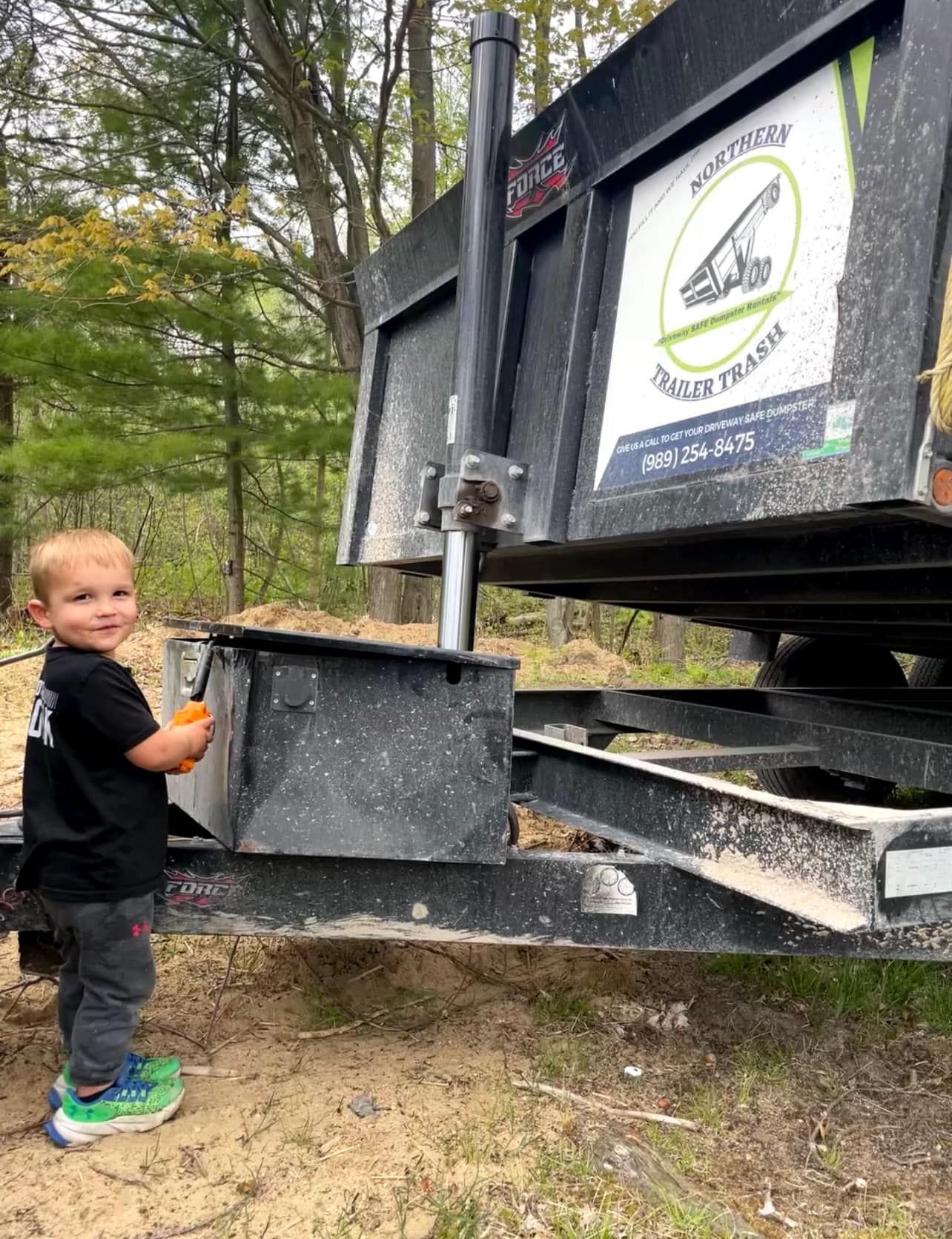 Child next to a dumpster. The child holds something orange, smiling. Green trees in the background.
