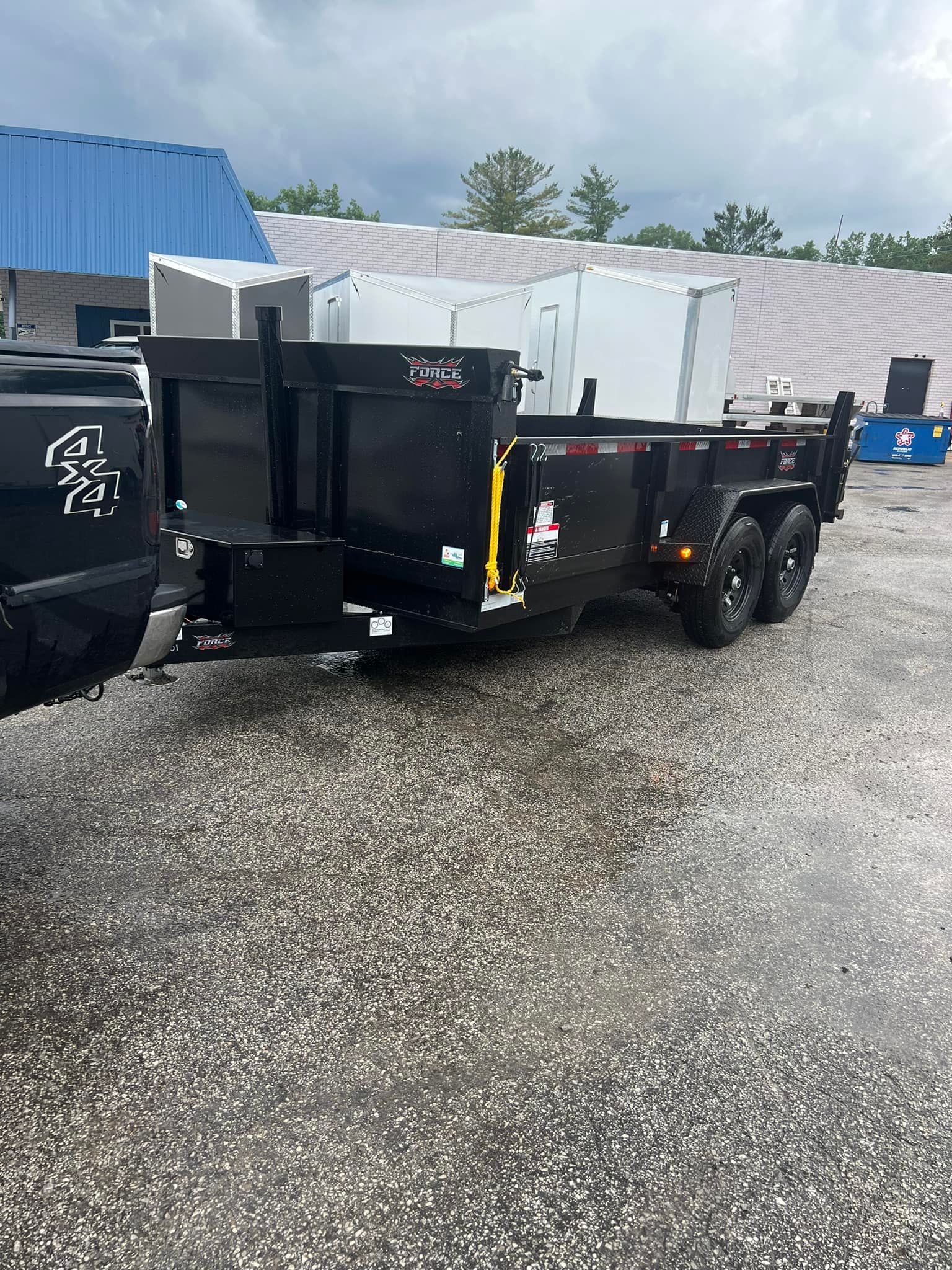 Black dump trailer hitched to a truck, parked on a paved lot.