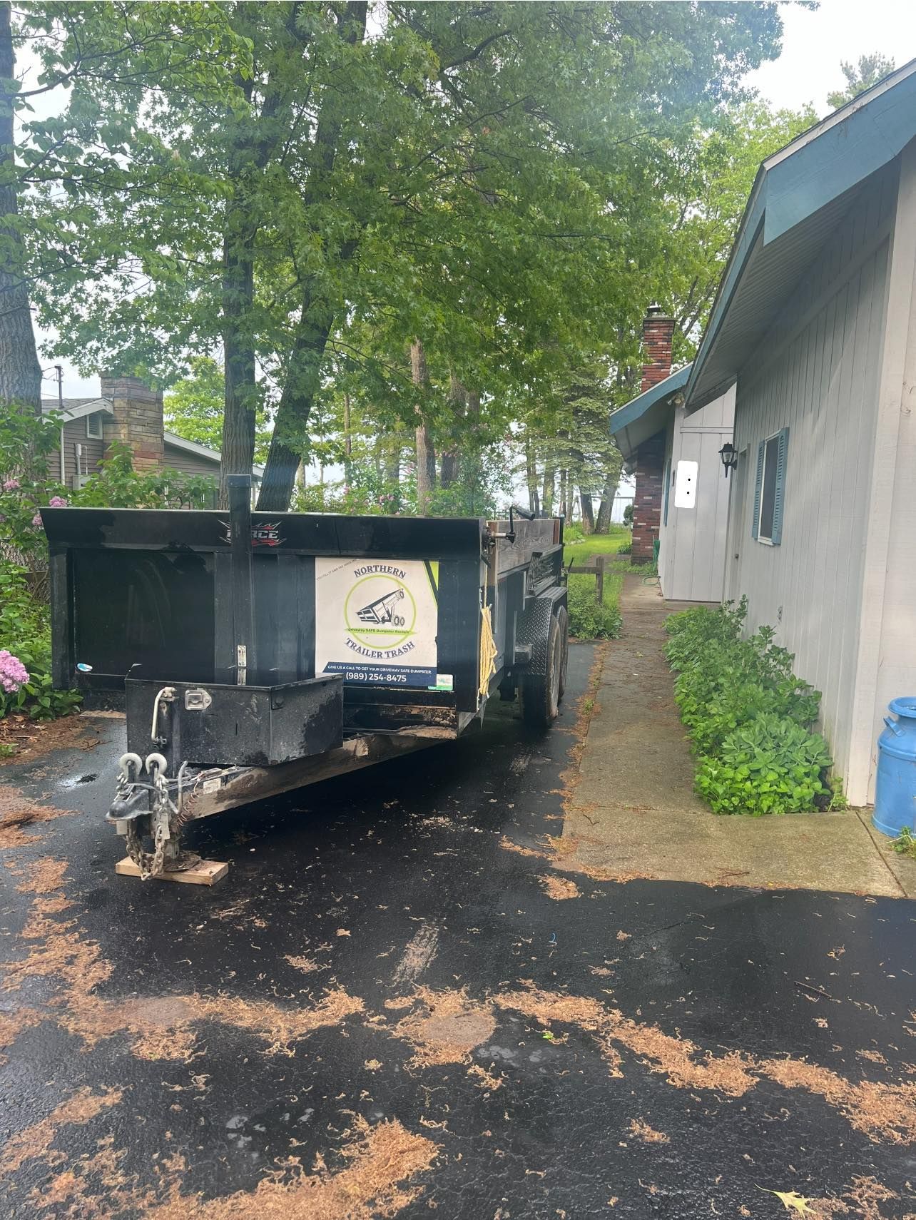 Black dumpster trailer parked next to a light-colored building with a narrow pathway and trees in the background.