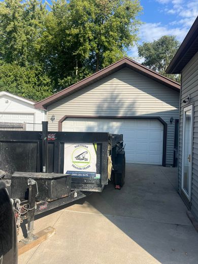 A black trailer backs toward a light beige garage with a white door. A gray house is on the right.