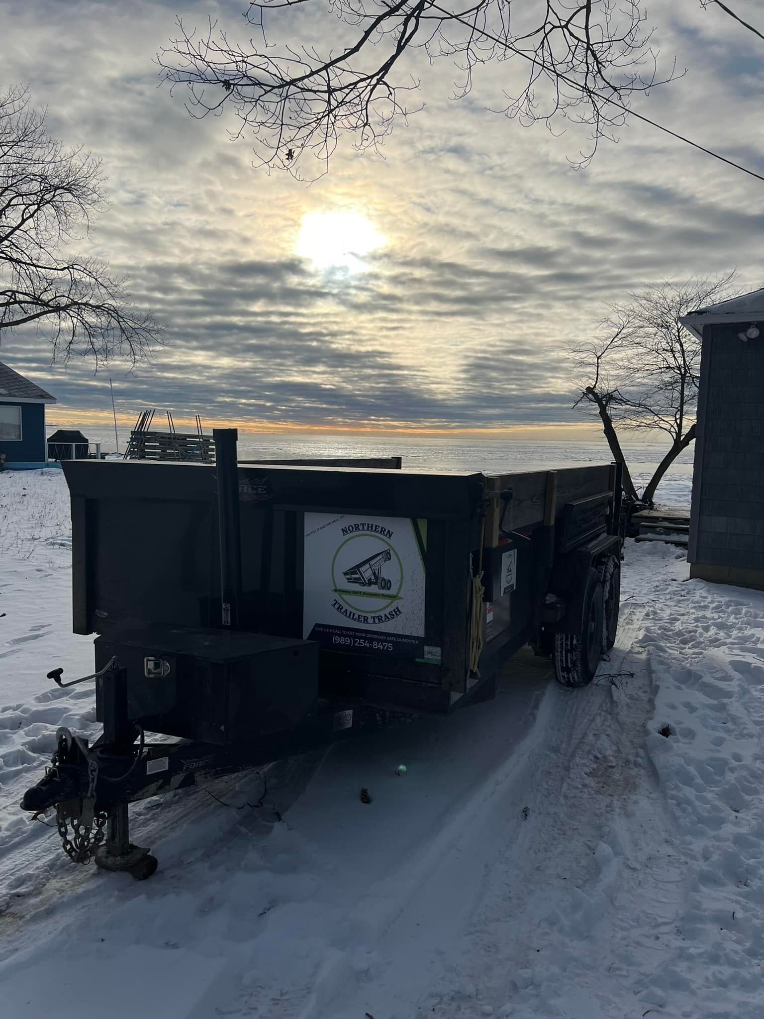 Black dump trailer on snowy ground with cloudy sky and water in the background.