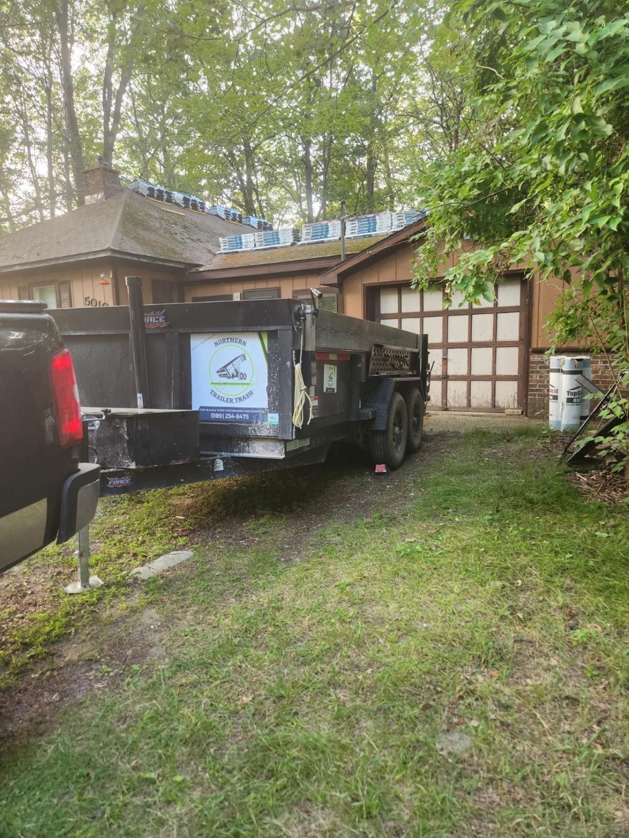 Trailer loaded with debris in front of a house. Roofing materials visible on the house.