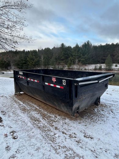 Black dumpster in a snowy outdoor setting with trees in the background.