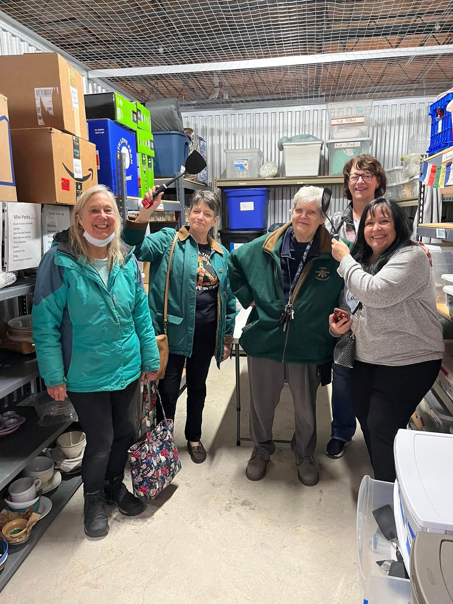 Four people in teal jackets standing in a storage room, smiling at the camera.