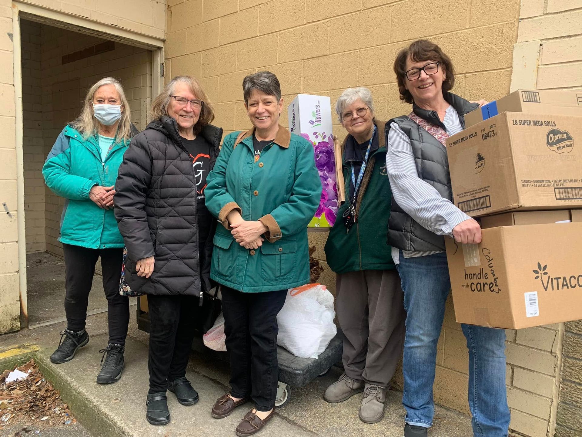 Five volunteers stand outside a building beside boxes of donated goods.