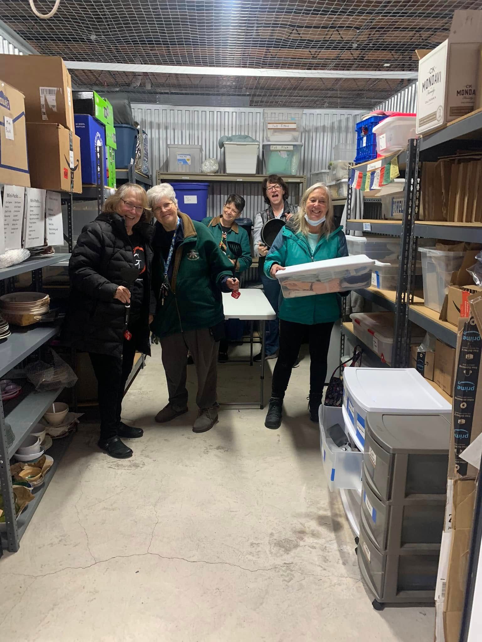People standing in a narrow storage aisle holding boxes and supplies, smiling at the camera.