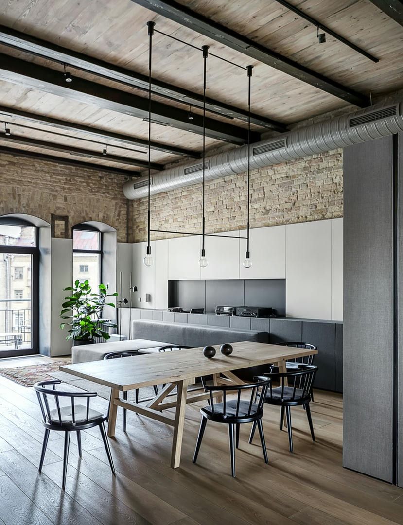 Industrial-style dining room with exposed brick, wood floor, and modern kitchen. Light fixtures hang over the wooden table and chairs.