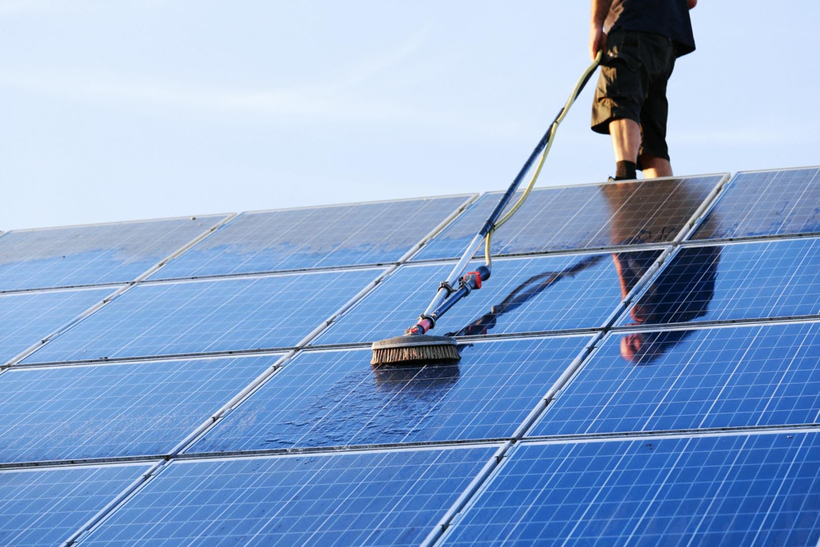 Person cleaning solar panels on a roof with a long-handled brush, blue panels, bright sunlight.
