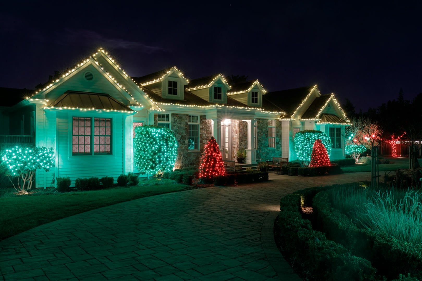 House decorated with green and white Christmas lights. Red tree and bushes on the lawn at night.