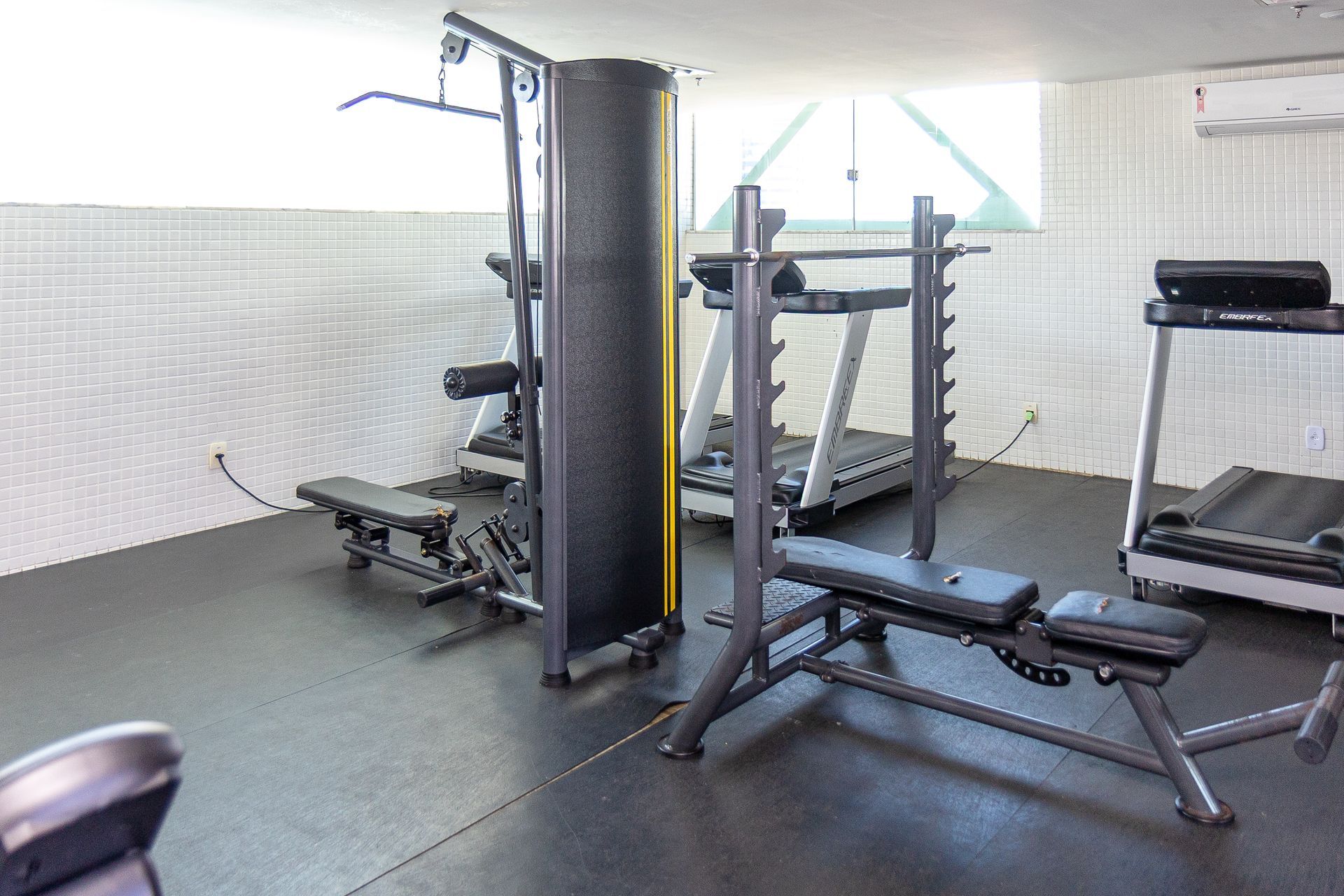 Sala de ginásio pequena, com máquinas de musculação, bancos e piso cinzento junto a uma janela