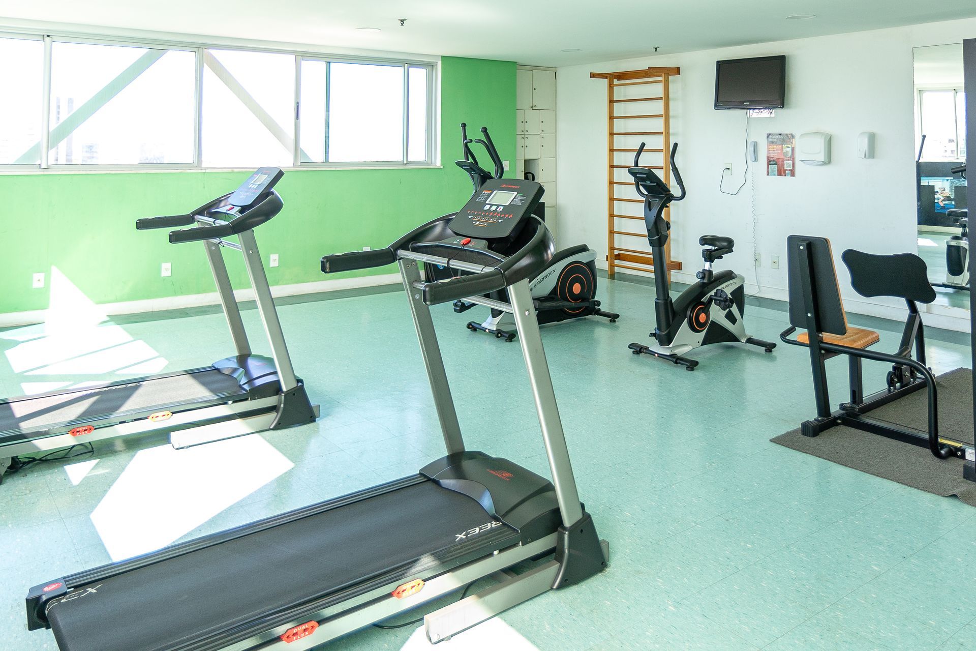 Sala de ginástica bem iluminada, com passadeiras, bicicletas ergométricas e aparelhos de musculação sobre um piso verde
