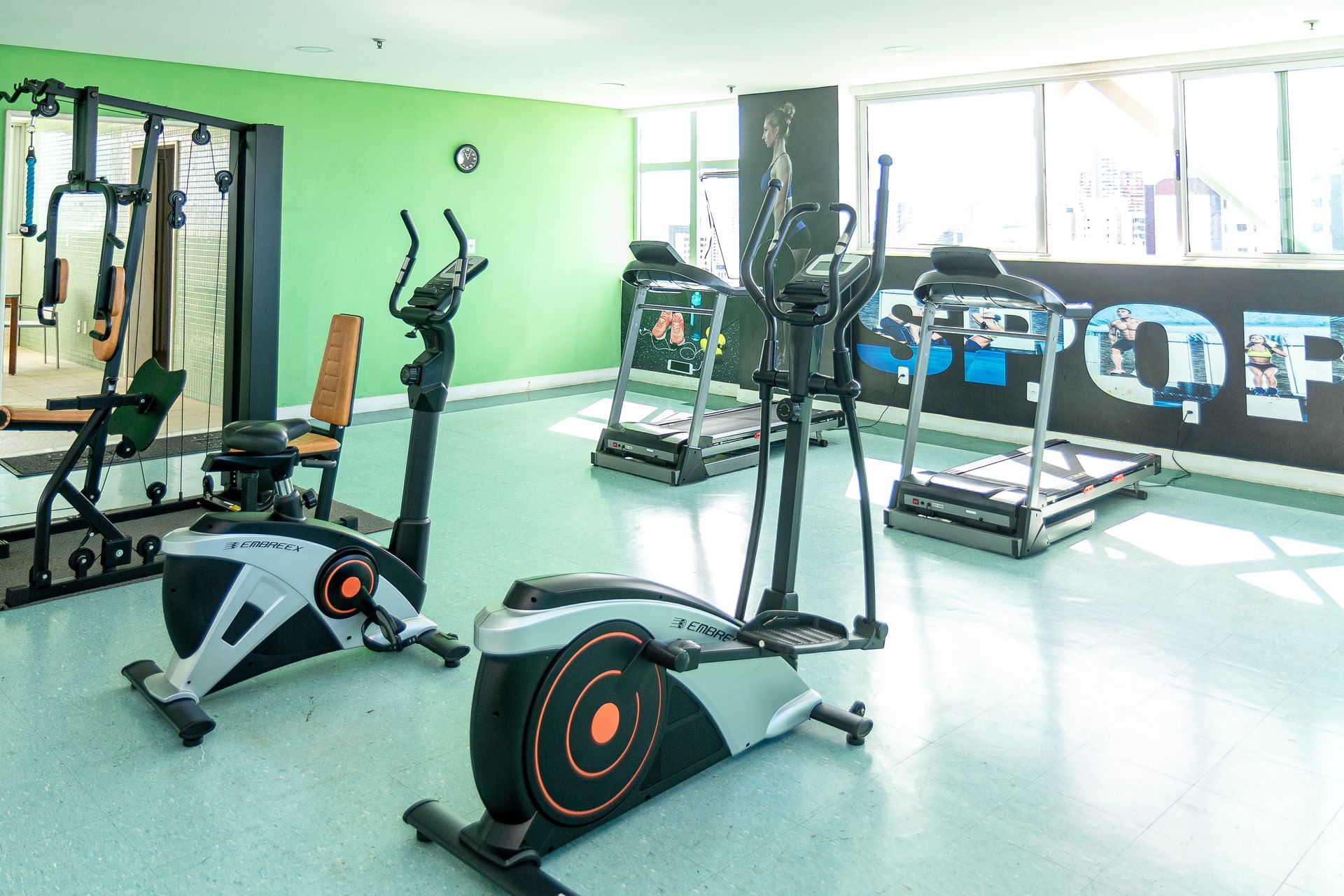 Academia bem iluminada, com aparelhos de cardio e equipamentos de musculação sobre um piso e diante de uma parede verdes.