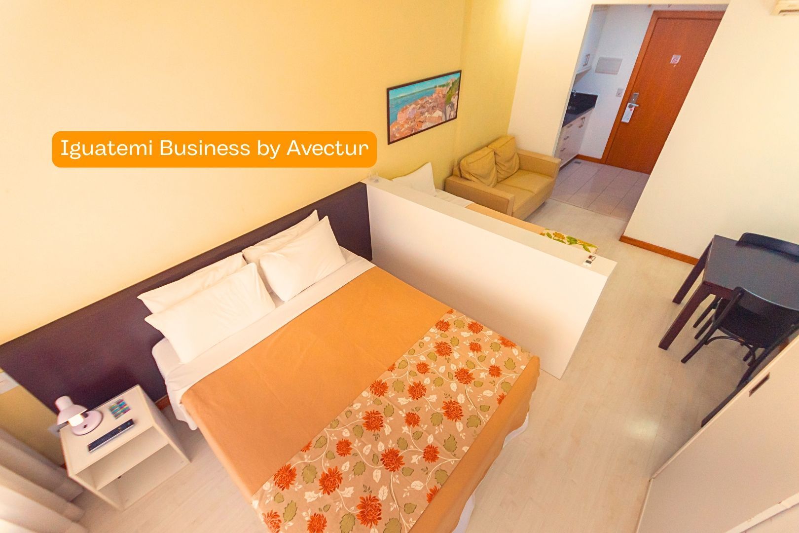 Quarto pequeno de hotel com duas camas, paredes brancas, detalhes em laranja e uma escrivaninha perto da porta.